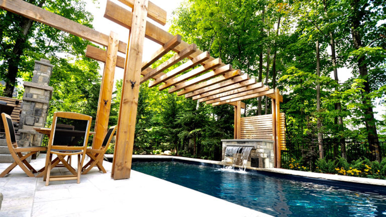 Stone pool deck teak table chairs beneath pergola. Lush forest in background. timber pergola spans across middle of swimming pool.