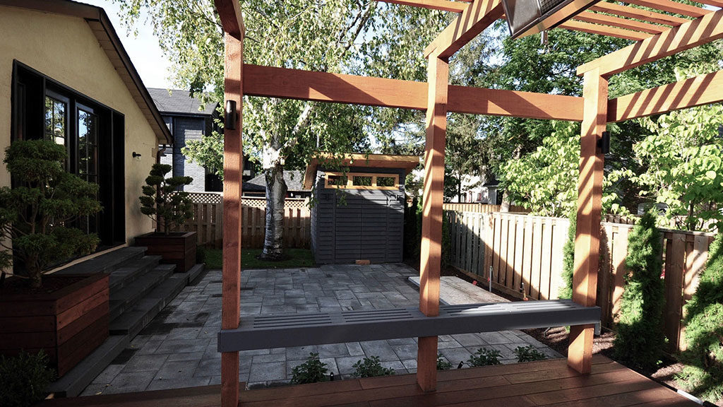 A view from deck through pergola posts to a walkout patio with a modern shed roof shed in the back corner.